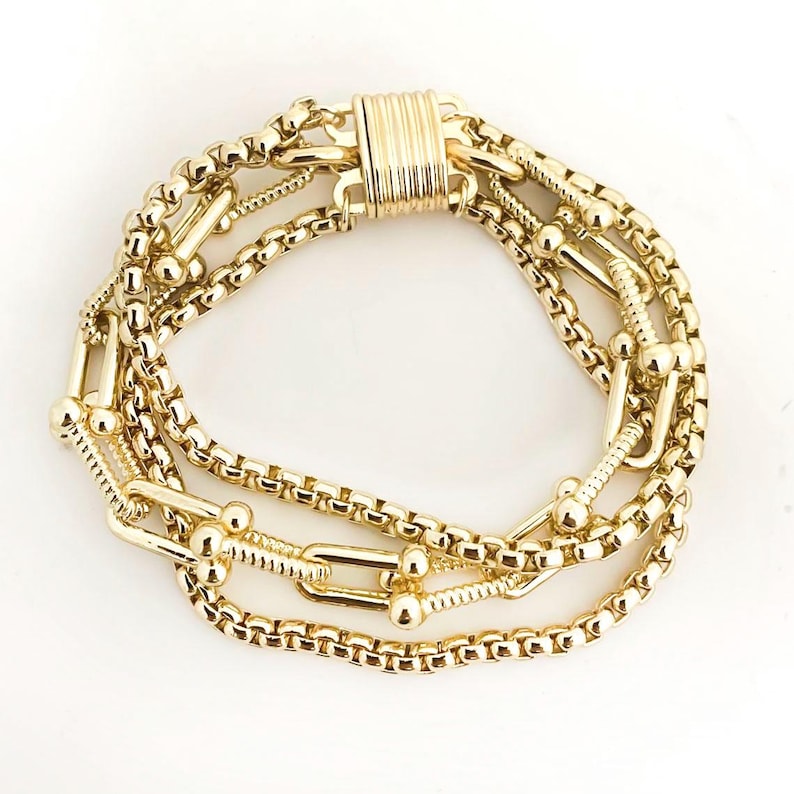 K&ouml;nnte beinhalten: Ein goldfarbenes Armband mit mehreren Str&auml;ngen verschiedener Kettenarten. Das Armband umfasst Boxketten, Kugelkettchen und einen Magnetverschluss. Der Schmuck wird auf wei&szlig;em Hintergrund pr&auml;sentiert.