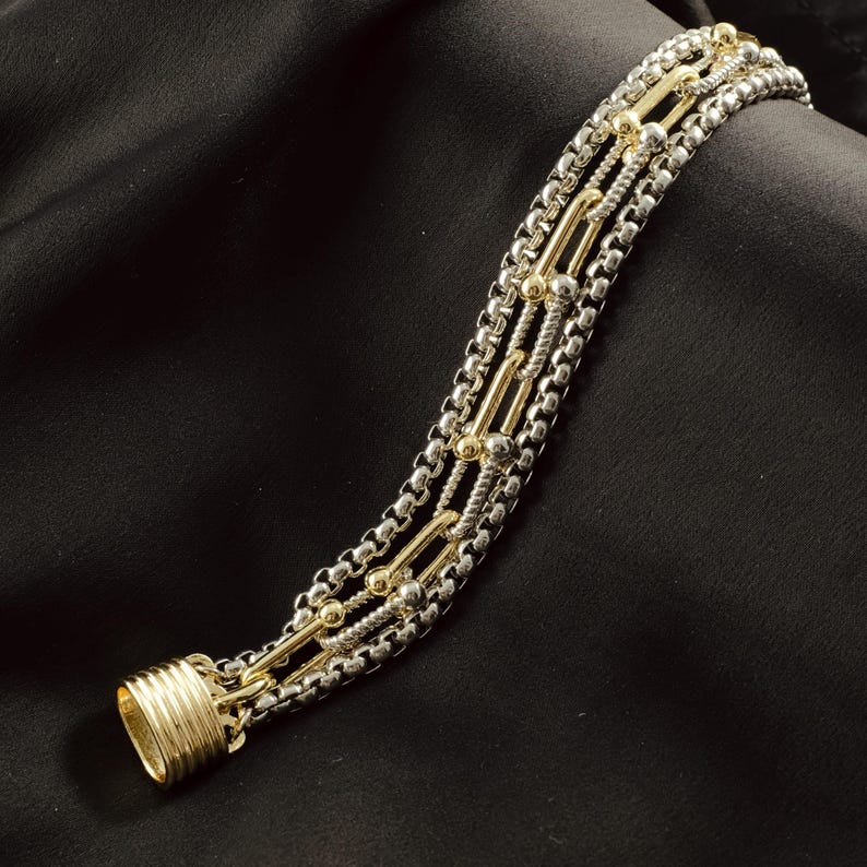 K&ouml;nnte beinhalten: Ein mehrreihiges Armband in Silber- und Goldt&ouml;nen. Das Armband hat eine Mischung aus Kettenstilen, darunter Boxketten und St&auml;be, mit einem goldfarbenen Verschluss. Der Schmuck wird auf einem schwarzen Hintergrund pr&auml;sentiert.