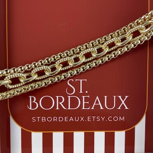 May include: Close-up of three gold-colored chain necklaces. The chains are layered and have different link styles. The background is a deep red with the text "ST. BORDEAUX" and the Etsy shop URL.