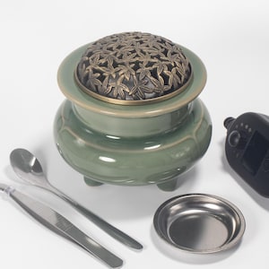 May include: A sage green ceramic incense burner with a bronze-colored, leaf-patterned lid. The burner has a rounded shape and sits on three small feet. Included are a small metal dish, tweezers, and a spoon.