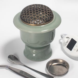Può includere: Un bruciatore di incenso in ceramica verde chiaro con un coperchio a rete metallica color bronzo. Il bruciatore ha una base ampia e piatta e un corpo arrotondato. Sono visibili anche un piccolo piatto di metallo, un cucchiaio e un pannello di controllo.