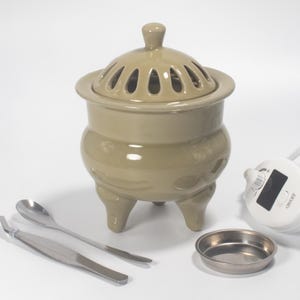 May include: A beige ceramic incense burner with a lid featuring oval-shaped vents. The burner has three small feet and is accompanied by a small metal dish, tweezers, a spoon, and a control panel.