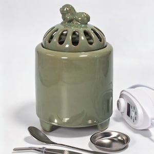 May include: A sage green ceramic incense burner with a cylindrical body and three short feet. The lid features a sculpted lion figure and oval cutouts. Included are a small metal tray, a metal spoon, metal tweezers, and a white power controller.