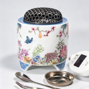 May include: A cylindrical white ceramic incense burner featuring a colorful floral and butterfly pattern, topped with a black metal lattice lid. It stands on three small feet and is accompanied by a metal tray, a spoon, and a white electronic control unit.