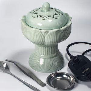 May include: A celadon green ceramic incense burner with a lotus flower design. The burner has a lid with decorative cutouts and sits on a pedestal base. Included are a small metal dish, a spoon, tweezers, and a control unit with an on/off switch.