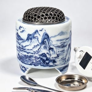 Può includere: Un bruciatore di incenso cilindrico in ceramica bianca con un dipinto paesaggistico blu di montagne e alberi. Ha un coperchio a cupola in rete metallica scura e poggia su tre piedini. Include un vassoio in metallo, un cucchiaio in metallo e un controller elettrico bianco.