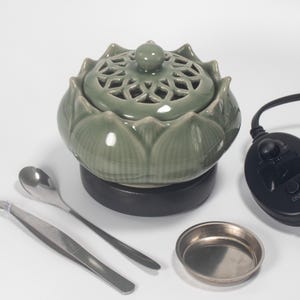 May include: A sage green ceramic lotus flower-shaped incense burner with a perforated lid. Includes a black base, a small metal dish, a spoon, tweezers, and a control panel. The burner is designed for aromatherapy and home decor.