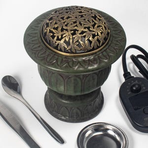 May include: Green ceramic incense burner with a decorative bronze lid featuring a leaf pattern. Includes a spoon, tweezers, a small metal dish, and a control unit with a digital display. The burner has a detailed, textured design.