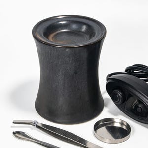Może przedstawiać: Czarny ceramiczny podgrzewacz do wosku o cylindrycznym, klepsydrowym kształcie z wgłębieniem na górze. W zestawie znajduje się mała srebrna metalowa miseczka, srebrna metalowa pęseta, metalowa szpatułka oraz czarny przewód zasilający z pokrętłem.
