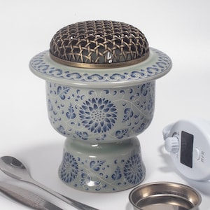 May include: A ceramic incense burner with a blue and white floral pattern. The burner has a woven metal lid and a small, round base. A spoon, tweezers, and a small metal bowl are next to the burner.