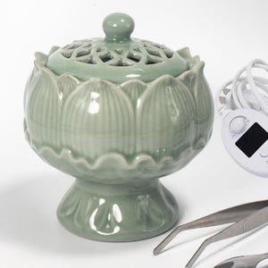 May include: A celadon green ceramic incense burner with a lotus flower design. The lid has a decorative cutout pattern. The burner sits on a pedestal base. A white control panel and metal tweezers are in the background.