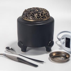 May include: A black ceramic incense burner with a decorative bronze-colored lid featuring a floral design. The burner has three small feet and comes with a spoon, tweezers, and a small metal dish. A white device with a dial is also present.