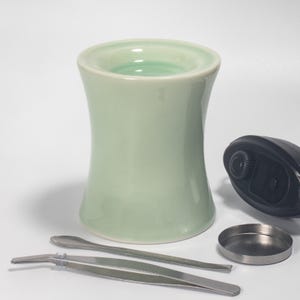 May include: A light green ceramic container with a flared top and base, accompanied by a black device, a small metal dish, a spoon, and tweezers. The container has a smooth, glossy finish. The items are arranged on a white surface.