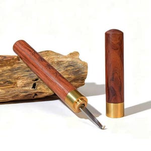 Op de afbeelding: Een houtsnijgereedschap met een cilindrische roodbruine houten handgreep en een messing huls. Het gereedschap heeft een gebogen metalen mes dat uit de handgreep steekt. Een bijpassende cilindrische houten dop met messing voet staat ernaast.