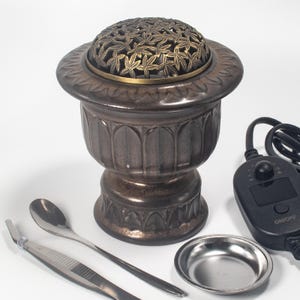 May include: A bronze-colored incense burner with a decorative leaf-patterned lid. The burner has a pedestal base and a matching small dish, spoon, and tweezers. A black control panel with an on/off switch is also included.