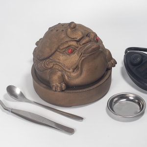 Puede incluir: Un quemador de incienso con forma de rana de color bronce con ojos rojos. El quemador se encuentra sobre una base circular, acompañado de un pequeño plato de metal, una cuchara, unas pinzas y un temporizador negro. La rana tiene detalles y una superficie texturizada.