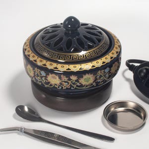 May include: A black ceramic incense burner with a decorative floral and geometric design in gold. The burner has a perforated lid and sits on a dark brown base. Accessories include a spoon, tweezers, and a small metal dish.