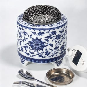 Può includere: Un bruciatore di incenso cilindrico in ceramica con motivo floreale blu e bianco, sormontato da una cupola in rete metallica scura. Sono inclusi un piccolo vassoio in metallo, un cucchiaio in metallo, pinzette in metallo e un'unità di controllo elettronica bianca con manopola.