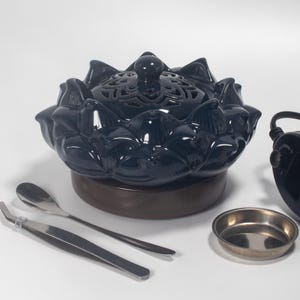 May include: A dark blue ceramic lotus flower-shaped incense burner with a decorative lid. The burner sits on a brown base, and is accompanied by a small metal dish, tweezers, and a spoon. The burner is designed for aromatherapy.