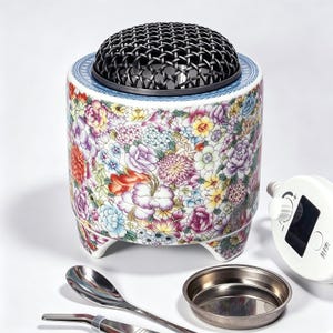 Pode incluir: Um queimador de incenso cilíndrico de cerâmica com um padrão floral colorido em tons de rosa, amarelo, roxo e laranja. Possui uma tampa de cúpula de malha metálica preta, uma pequena bandeja de metal, uma colher de metal e uma unidade de controle eletrônica branca com texto.