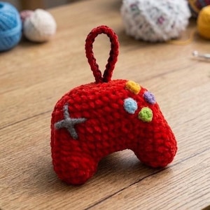 Peut inclure: Une manette de jeu vidéo rouge au crochet faite à la main, avec des boutons colorés et une croix directionnelle grise. La manette a une boucle pour l'accrocher. Du fil et du matériel de bricolage sont en arrière-plan.