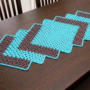 May include: A handmade crochet table runner in a zig-zag pattern. The runner features a turquoise and brown color scheme. The crochet design is a series of squares, creating a decorative accent for a dining table or other surface.
