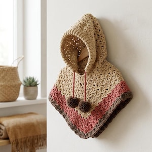 May include: A handmade, hooded poncho in beige, pink, and brown tones. The crochet poncho features a hood with a drawstring and two brown pom-poms. The poncho is hanging on a wooden hanger against a white wall.