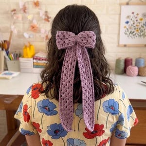 Op de afbeelding: Een lavendelkleurige gehaakte strik haaraccessoire met lange, vloeiende staarten. De strik is bevestigd aan de achterkant van het hoofd van een persoon en de staarten vallen naar beneden. De persoon draagt een gele bloemenjurk.