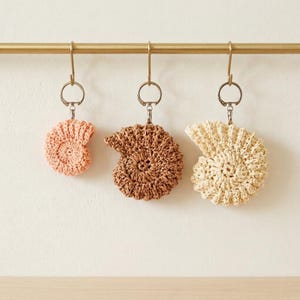 May include: Three crochet seashell keyrings in peach, brown, and cream hang from a gold bar. Each keyring features a spiral design and a silver-coloured ring. The keyrings are handmade and suitable for beach-themed decor.