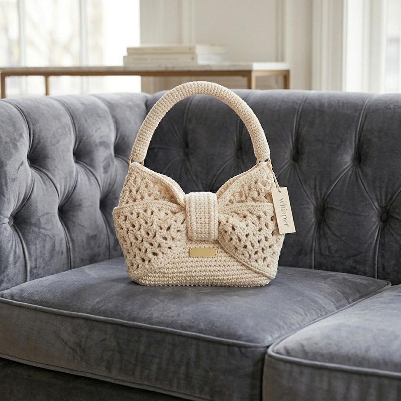 Peut inclure: Un sac &agrave; main en crochet cr&egrave;me avec un devant en forme de n&oelig;ud papillon et une poign&eacute;e arrondie. Le sac a un design en tissage ouvert et une petite &eacute;tiquette rectangulaire avec le mot "whisper". Le sac est pos&eacute; sur un canap&eacute; gris.