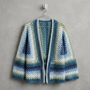 May include: A crocheted cardigan in shades of blue, green, and white hangs on a metal hanger. The open-front sweater features a geometric pattern and long sleeves. The garment is displayed against a neutral grey wall.
