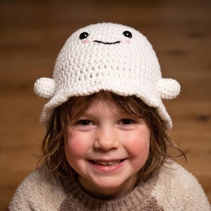 Può includere: Un cappello a secchiello all'uncinetto bianco con un design a fantasma, due piccole sporgenze laterali, occhi ricamati neri e una bocca cucita nera. Il cappello presenta delicati tocchi di rosa sotto gli occhi, creando un aspetto rotondo e amichevole.