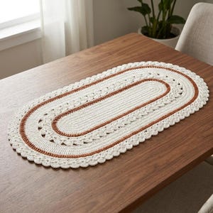 May include: Oval, cream-coloured crochet table runner with a brown border. The runner is placed on a wooden table. The crochet design features a scalloped edge and a central oval pattern. The runner is approximately 61 cm long.