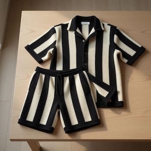May include: A matching two-piece set of black and cream-colored striped clothing. The set includes a short-sleeved button-down shirt and a pair of shorts with a drawstring waist. The fabric has a textured waffle-knit appearance, and the set is displayed on a light-colored wooden surface.