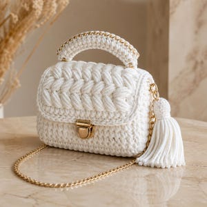 Peut inclure: Un sac à main blanc, crocheté à la main, avec un motif tressé sur le rabat avant. Le sac est doté d'une chaîne et d'une poignée dorées, d'un fermoir doré et d'un pompon blanc. Le sac est posé sur une surface en marbre.