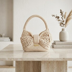 Peut inclure: Un sac &agrave; main en crochet beige avec un rabat avant en forme de n&oelig;ud papillon et une poign&eacute;e sup&eacute;rieure. Le sac pr&eacute;sente un design ajour&eacute; textur&eacute; et une petite plaque rectangulaire dor&eacute;e. Une &eacute;tiquette avec le mot "whisper" est suspendue &agrave; la poign&eacute;e.