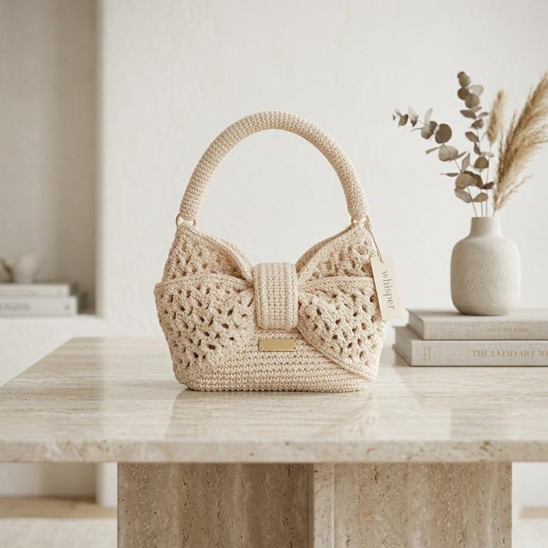Peut inclure: Un sac &agrave; main en crochet beige avec un rabat avant en forme de n&oelig;ud papillon et une poign&eacute;e sup&eacute;rieure. Le sac pr&eacute;sente un design ajour&eacute; textur&eacute; et une petite plaque rectangulaire dor&eacute;e. Une &eacute;tiquette avec le mot "whisper" est suspendue &agrave; la poign&eacute;e.