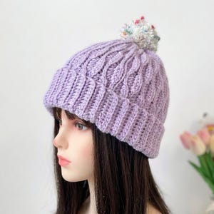 May include: A lilac knitted beanie hat with a cable knit pattern and a ribbed brim. The hat is topped with a fluffy, multi-coloured pom-pom. This winter hat is a warm accessory.