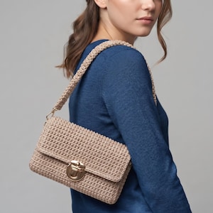 May include: A beige crochet shoulder bag featuring a rectangular silhouette with a textured, knotted stitch pattern. The bag includes a matching thick crochet strap and a gold-toned metal push-lock clasp closure centered on the front flap.