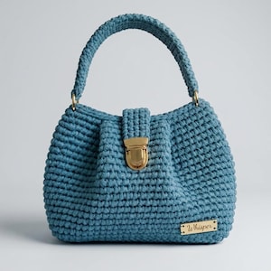 May include: A blue crochet handbag with a rounded shape and a top handle. The bag features a gold-colored clasp and hardware. A small gold-colored rectangular tag with the word "Whisper" is attached to the front of the bag.