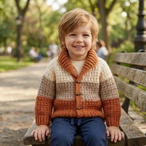 Puede incluir: Un niño lleva una rebeca de ganchillo hecha a mano. La rebeca tiene cuello de solapa y botones. La combinación de colores incluye crema, marrón y naranja. El niño está sentado en un banco de madera en un parque, con la rebeca puesta.