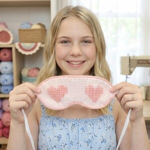 May include: A handmade crochet sleep mask featuring a white base with two pink heart patterns in the center. The mask has a light pink crocheted border and a thin white elastic strap attached to each side for securing it around the head.