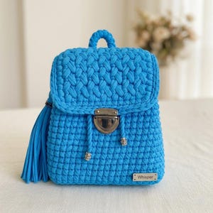 Può includere: Uno zaino all'uncinetto blu brillante con chiusura a patta e chiusura argentata. La borsa presenta un manico superiore intrecciato, una lunga nappa e dettagli decorativi argentati. La parola "Whisper" è su una piccola etichetta argentata.