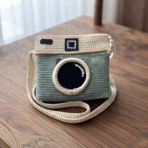 May include: A crochet bag shaped like a vintage camera. The body is sage green with a cream-colored top and strap. A large, circular black lens sits in the center, surrounded by a cream border. Small dark rectangular details accent the top section.