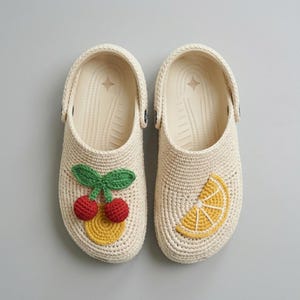 May include: A pair of cream-colored clogs with crochet detailing. One clog features a cherry design with red cherries and green leaves, while the other has a yellow lemon slice. The clogs have a rubber sole.