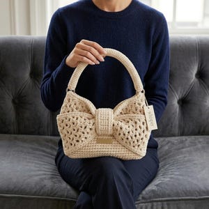 Peut inclure: Un sac &agrave; main en crochet beige avec un motif en forme de n&oelig;ud. Le sac a une forme structur&eacute;e avec une poign&eacute;e et une petite &eacute;tiquette qui dit "whisper". Le sac est tenu par une personne portant un pull bleu marine.