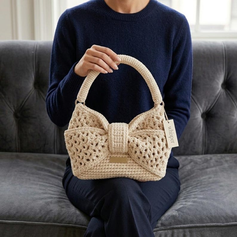 Peut inclure: Un sac &agrave; main en crochet beige avec un motif en forme de n&oelig;ud. Le sac a une forme structur&eacute;e avec une poign&eacute;e et une petite &eacute;tiquette qui dit "whisper". Le sac est tenu par une personne portant un pull bleu marine.