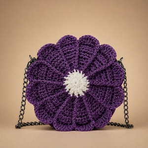 May include: A handmade, purple crochet bag shaped like a flower with a white center. The bag has a black chain strap. The flower petals are detailed with textured stitching, and the bag is set against a neutral background.