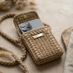 Op de afbeelding: Een beige gehaakte telefoon crossbody tas met een klepsluiting en een goudkleurige metalen plaat. De tas heeft een gevlochten band en er is een telefoon zichtbaar. De tas is gemaakt van een getextureerd, geweven materiaal.