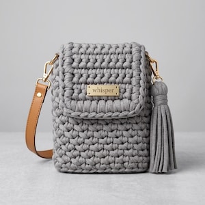 Op de afbeelding: Een grijze gehaakte crossbodytas met een bruine leren riem en goudkleurige hardware. De tas heeft een bijpassende grijze kwast en een klein goudkleurig naamplaatje met de tekst "whisper".